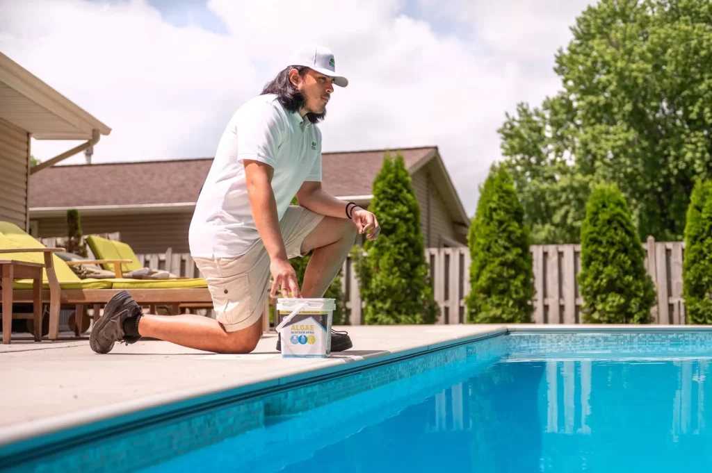 Pool technician kneeling beside pool with Algae Blocker bucket.